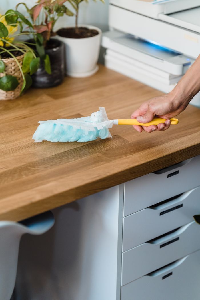 A hand with a feather duster dusting a wooden desk, with plants and office accessories nearby.
